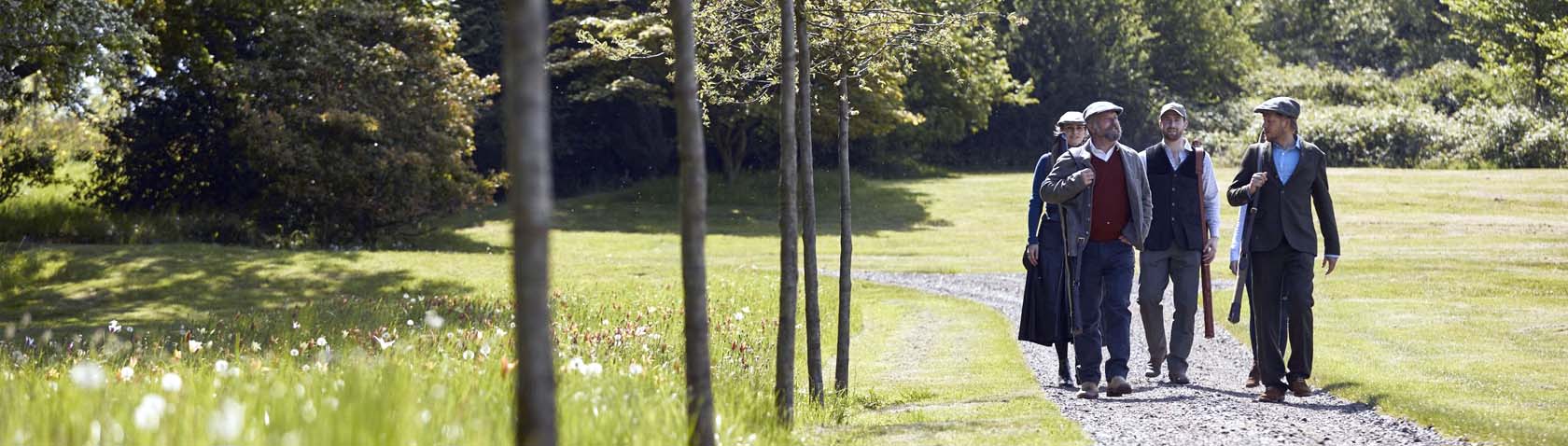 A group of well-dressed gentlemen walking along a scenic, tree-lined path in the countryside, surrounded by lush greenery and sunlight filtering through the trees.