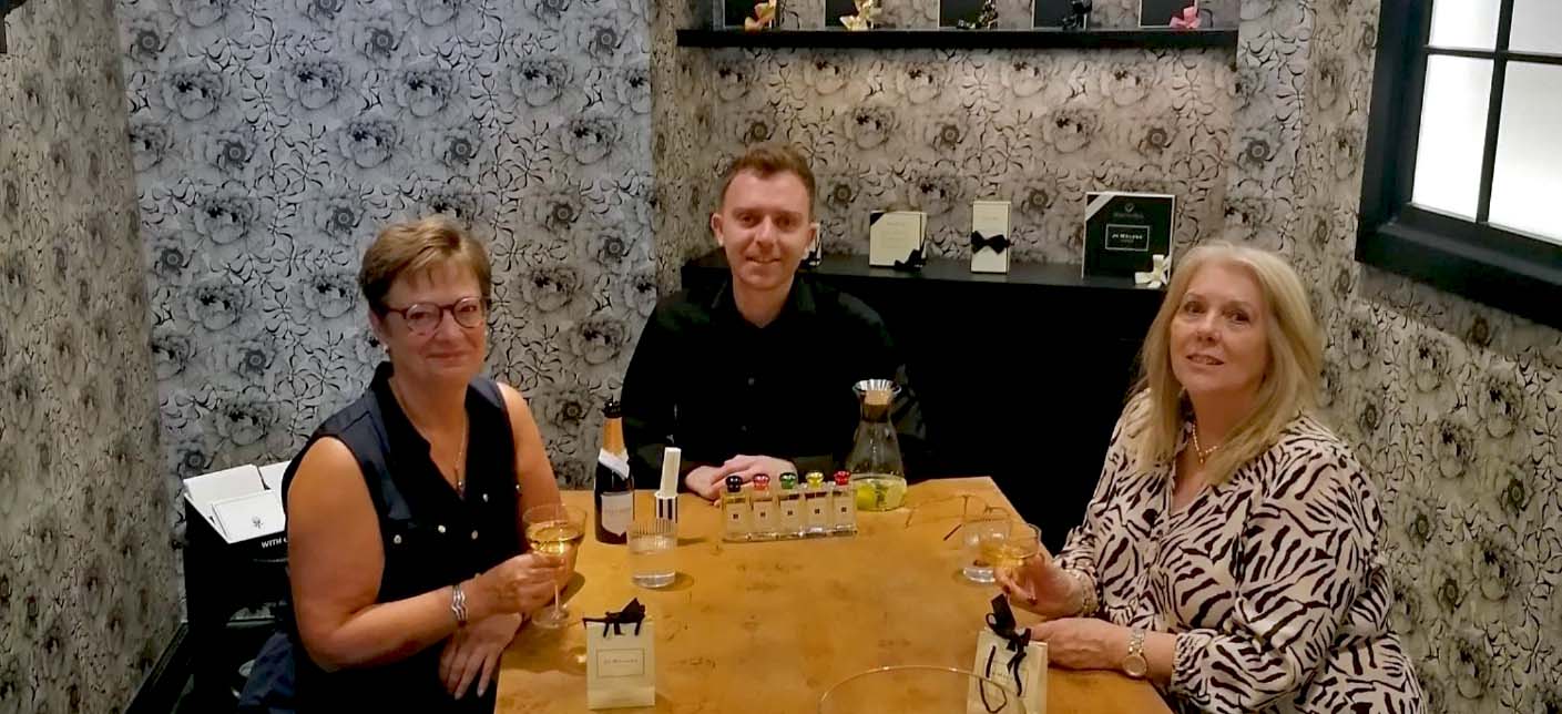 Three people enjoying a personalised fragrance experience at a Jo Malone perfume store, seated around a wooden table with perfume bottles and a bottle of champagne.