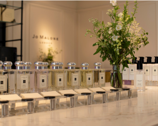 A refined display of Jo Malone perfume bottles arranged neatly on a marble counter with a floral arrangement in the background, showcasing a luxurious and elegant shopping environment.