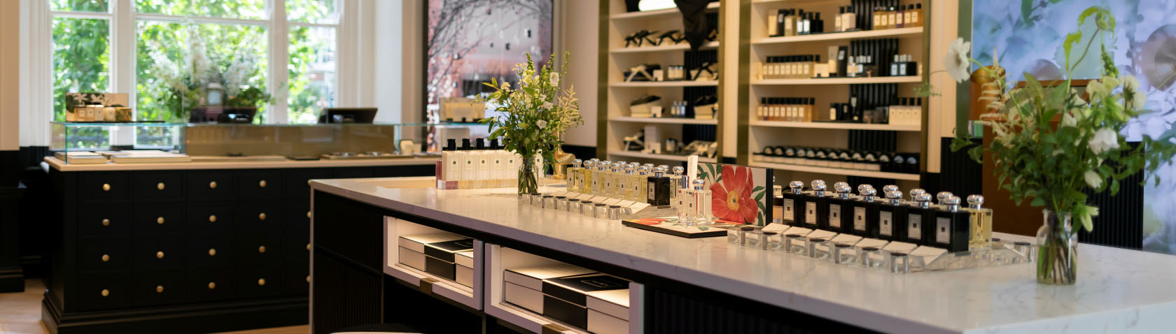 Interior view of a luxury perfume store showcasing elegant displays of various perfume bottles on a marble counter, surrounded by stylish shelving and floral arrangements.