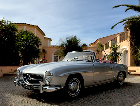 Classic silver Mercedes-Benz convertible parked in front of an elegant villa with palm trees.