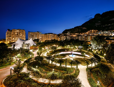 Nighttime view of a beautifully illuminated garden and fountain surrounded by high-rise buildings and mountains in Monte Carlo.