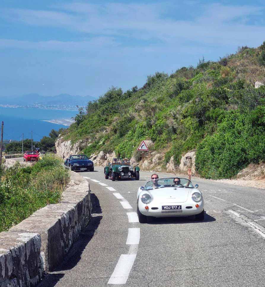 Scenic coastal road with vintage cars driving in a convoy, featuring lush greenery and a clear blue sky, with a distant view of the sea and mountains.