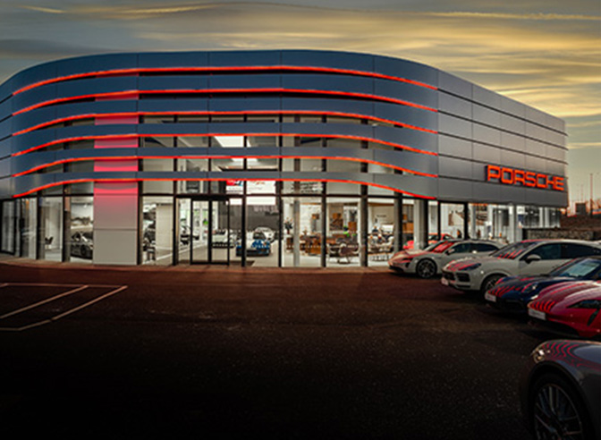 The exterior of a modern Porsche dealership at dusk. The building is characterised by its sleek, curved architecture with illuminated red accents. A lineup of various Porsche models is parked in front.
