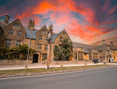 The Lygon Arms, a historic hotel in the Cotswolds, set against a dramatic orange sunset sky. The building is made of traditional Cotswold stone, featuring classic architecture with multiple gabled roofs and mullioned windows.