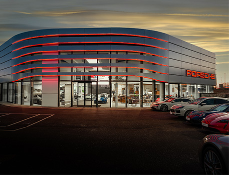 The exterior of a modern Porsche dealership at dusk. The building is characterised by its sleek, curved architecture with illuminated red accents. A lineup of various Porsche models is parked in front, showcasing the luxury and sportiness of the brand.