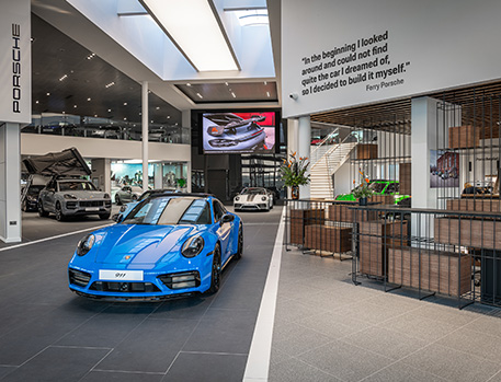 A modern Porsche dealership interior, featuring a blue Porsche 911 in the foreground and various other models. Overhead, skylights illuminate the showroom, which is adorned with a quote from Ferry Porsche and equipped with a large digital screen.