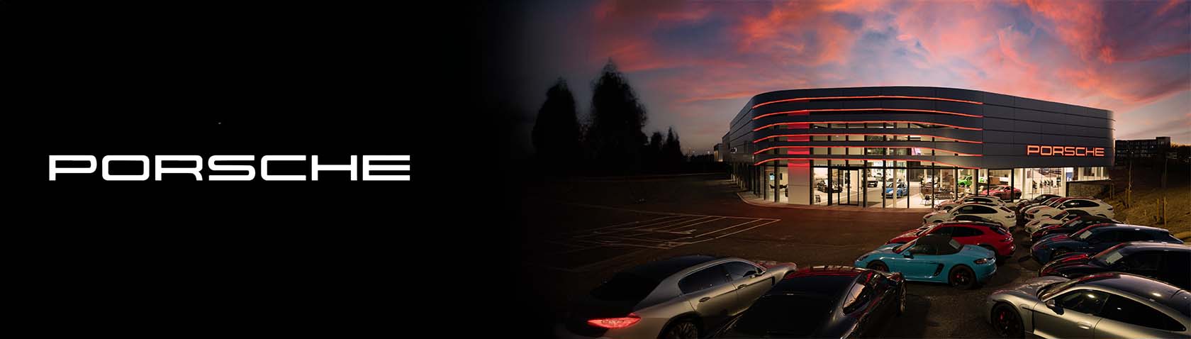 This image features a panoramic view of a Porsche dealership with a vibrant sky in the background. Various Porsche models are displayed in front of the modern, illuminated building.