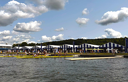 Rowing competition at Henley Royal Regatta with rowers and spectators near striped tents along the riverside on a sunny day.