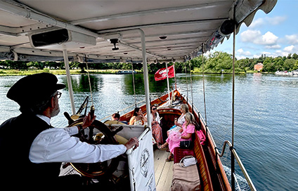 Captain steering a steam-powered boat with passengers enjoying the scenic view during the Henley Royal Regatta.