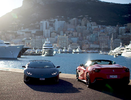 Two high-end sports cars, a dark-coloured Lamborghini and a red Ferrari, parked by the waterfront with a backdrop of Monaco’s cityscape and luxury yachts.