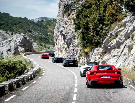 A convoy of luxury sports cars, including a prominent red Ferrari, driving on a winding road through a mountainous landscape.