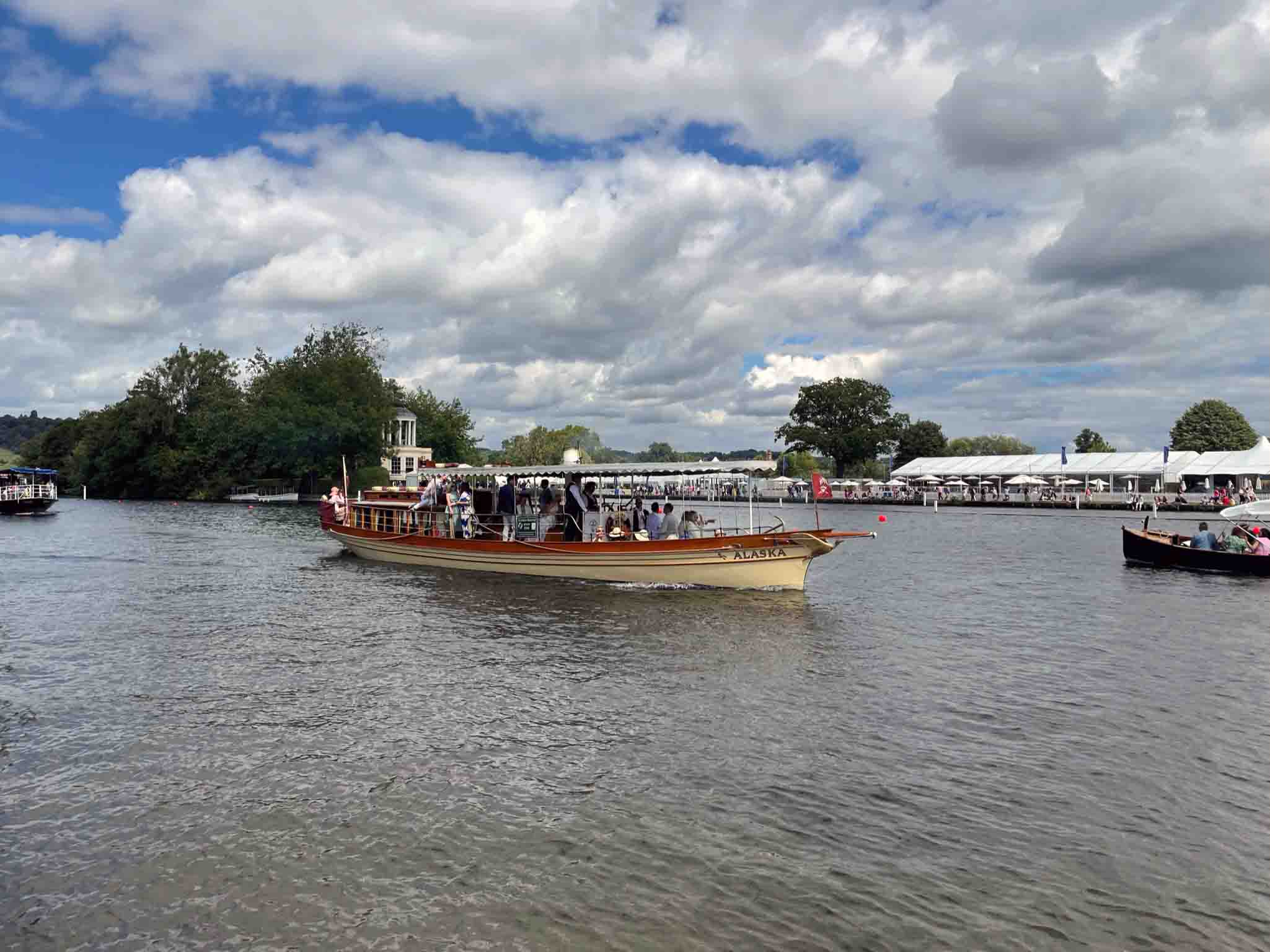 Steamboat Alaska cruising on the River Thames during the Henley Royal Regatta, with spectators and other boats in the background under a partly cloudy sky.