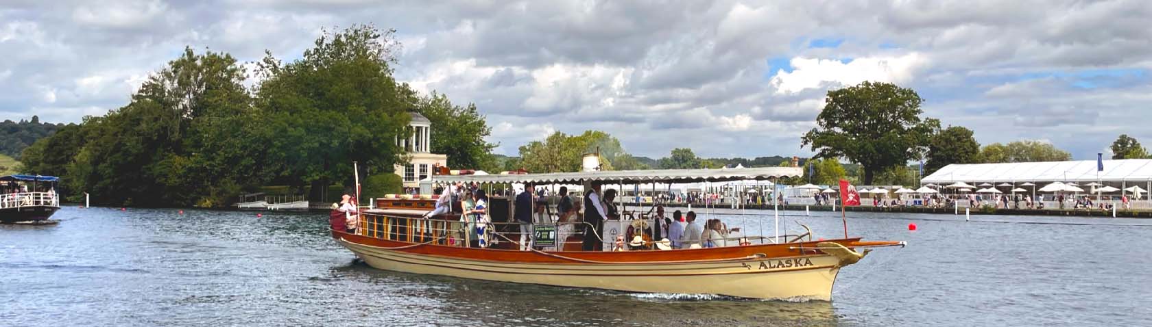 A vintage steamboat named "Alaska" cruising on the Thames River during the Henley Royal Regatta, with passengers enjoying the event, lush greenery, and event tents visible in the background.