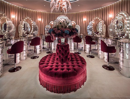 A luxurious makeup studio with velvet seating, ornate mirrors with lights, and elegant decor, centred around a plush round ottoman with a floral arrangement.