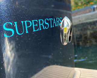 Close-up of the side of the Superstars private boat showing a polished metal cleat and the boat's sleek design, with water and greenery in the background.