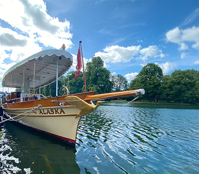 Vintage steamboat named "Alaska" docked on the Thames River, featuring an ornate bow design and shaded canopy.