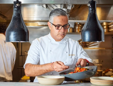 Chef in white uniform preparing a vibrant vegetable dish in a professional gourmet kitchen under pendant lights.