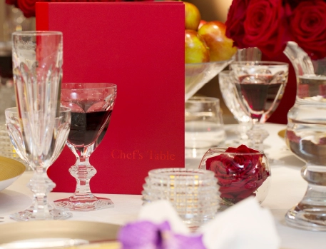 Elegant table setting at The Dorchester's Chef's Table, featuring crystal glasses, red wine, vibrant roses, and a refined red menu, creating a luxurious dining atmosphere.