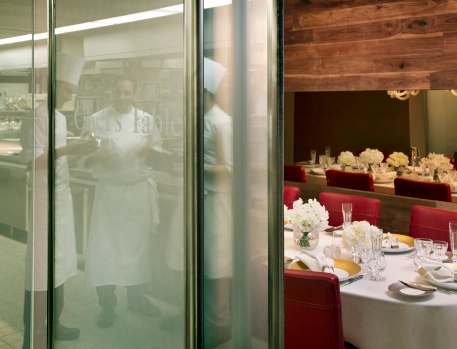 Chef's Table private dining room at The Dorchester, London, offering an exclusive view of the kitchen with chefs in preparation, featuring a refined table setting with white floral decor and red chairs.