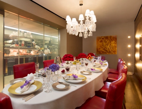 Luxurious Chef's Table private dining experience at The Dorchester, London, featuring a beautifully set table for a 5-course tasting menu with elegant red chairs, crystal chandeliers, and a view of the kitchen.