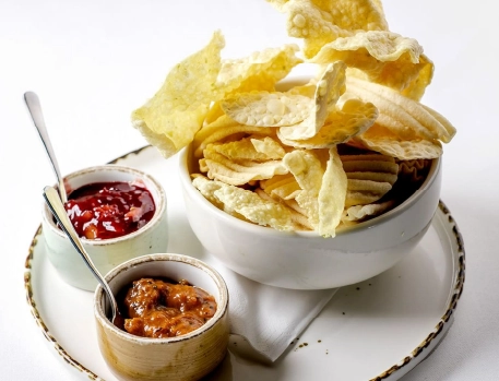 A bowl of assorted crispy pappadums served with two chutney dips on a white plate, perfect as an Indian starter.