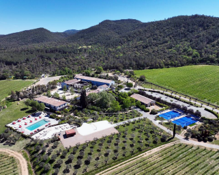 An aerial view of the Ultimate Provence Hotel nestled amidst lush vineyards and rolling hills. The property features a swimming pool with sun loungers, tennis courts, and various Mediterranean-style buildings surrounded by greenery.