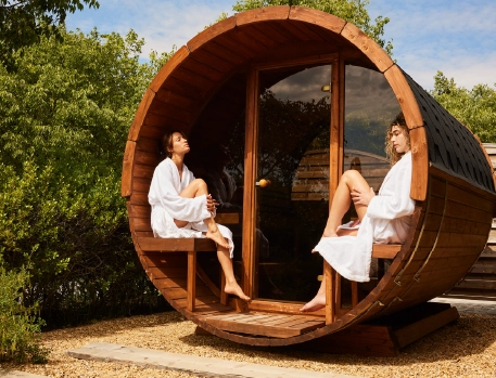 Two women relaxing in robes outside a wooden sauna barrel at Ultimate Provence Hotel, surrounded by greenery, enjoying a peaceful moment in the outdoor spa area.