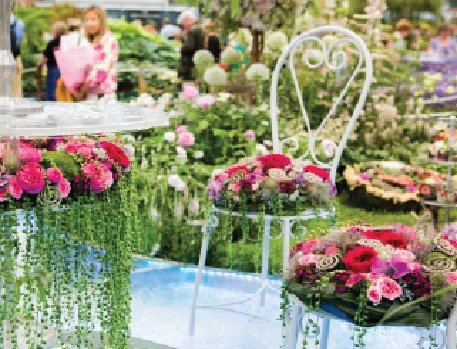 A beautifully arranged seating area with white wrought iron chairs and a table adorned with pink and red floral arrangements, set in a garden exhibition.